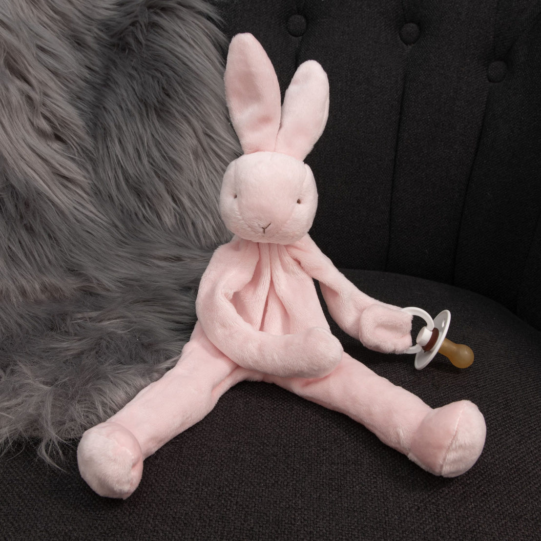 A Pink Silly Bunny Buddy, designed as a pacifier holder, rests on a dark cushioned chair. This charming baby gift gently holds a pacifier, with a fluffy gray fur blanket partially draped behind it.