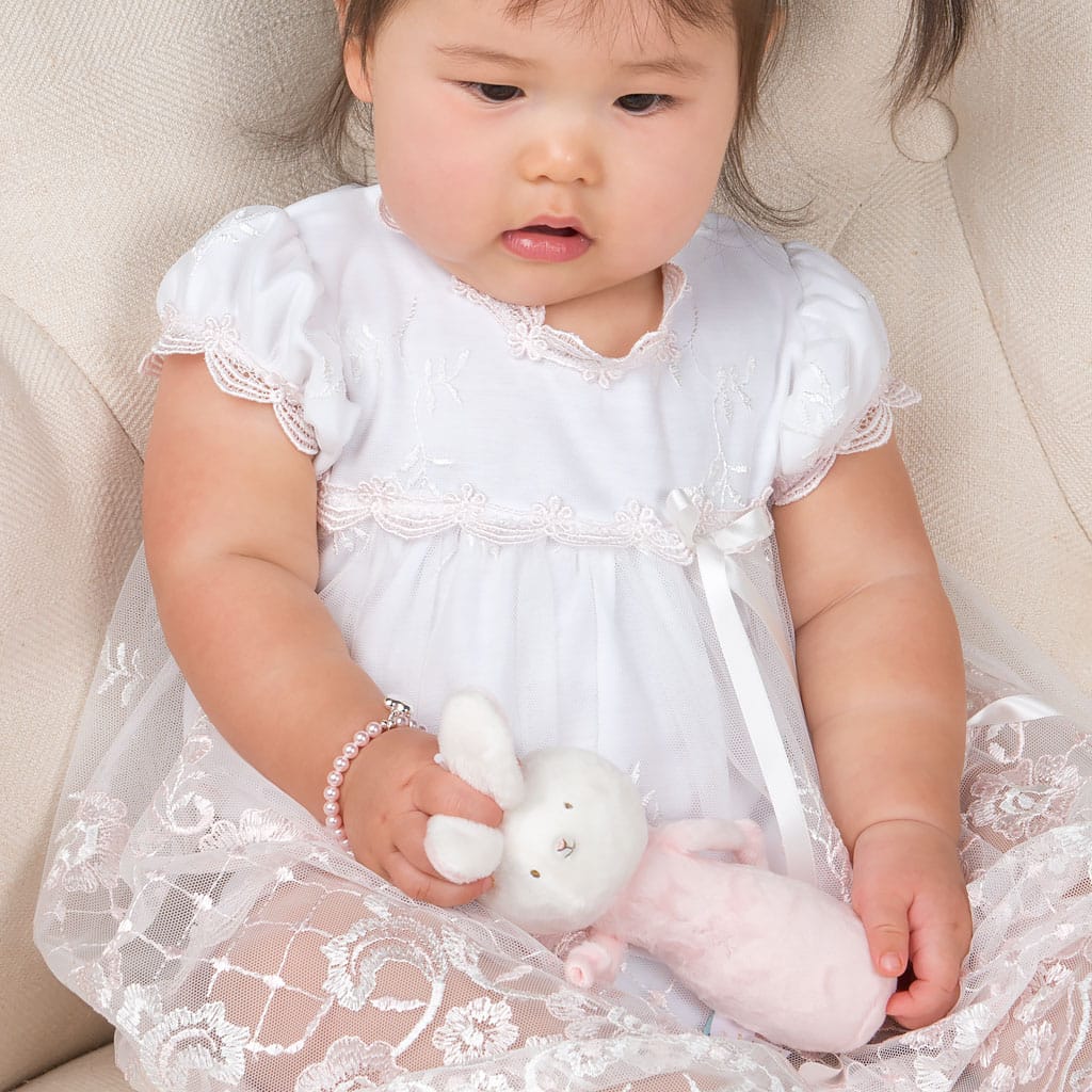 A toddler dressed in a white lace dress sits on a cream-colored chair, holding a Plush Chime Rattle. With dark hair and a bracelet on one wrist, the child gazes down at the baby-safe toy with a calm expression.