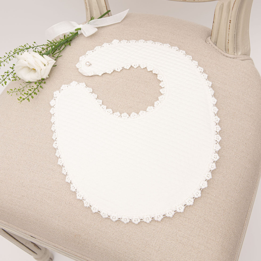 The Poppy Bib displayed on a beige chair, featuring quilted cotton fabric with scalloped lace trim, paired with a small white flower for a delicate presentation.