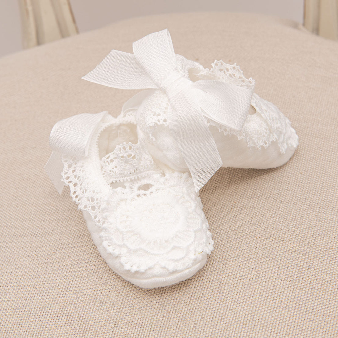 A pair of Poppy Booties displayed on a neutral textured chair, featuring delicate floral lace details and elegant white ribbon ties.