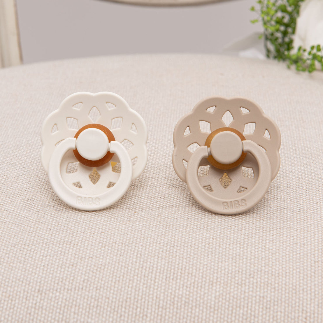 Scalloped pacifier set in vanilla and ivory tones, elegantly displayed on a soft beige surface with greenery detail nearby.