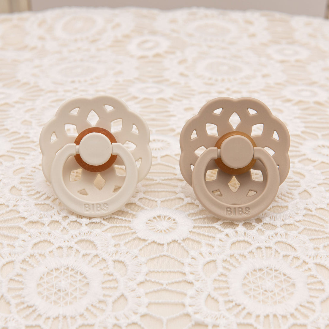 Pair of scalloped pacifiers in vanilla and ivory tones, displayed on a floral lace background, showcasing their delicate design.