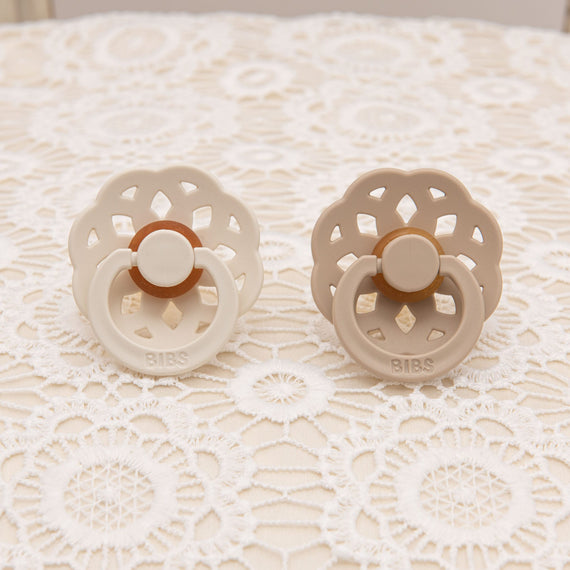 Pair of scalloped pacifiers in vanilla and ivory tones, displayed on a floral lace background, showcasing their delicate design.