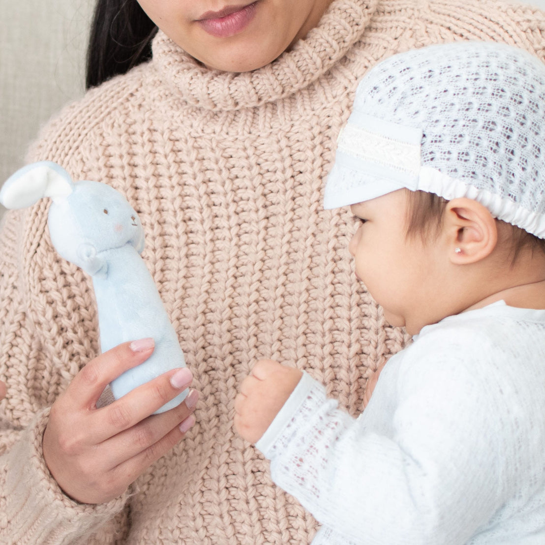 A mother in a beige sweater holds her baby during a baptism; the baby reaches for a Puppy Chime Rattle toy and wears a white knit hat, while only the lower half of the mother's face is visible.