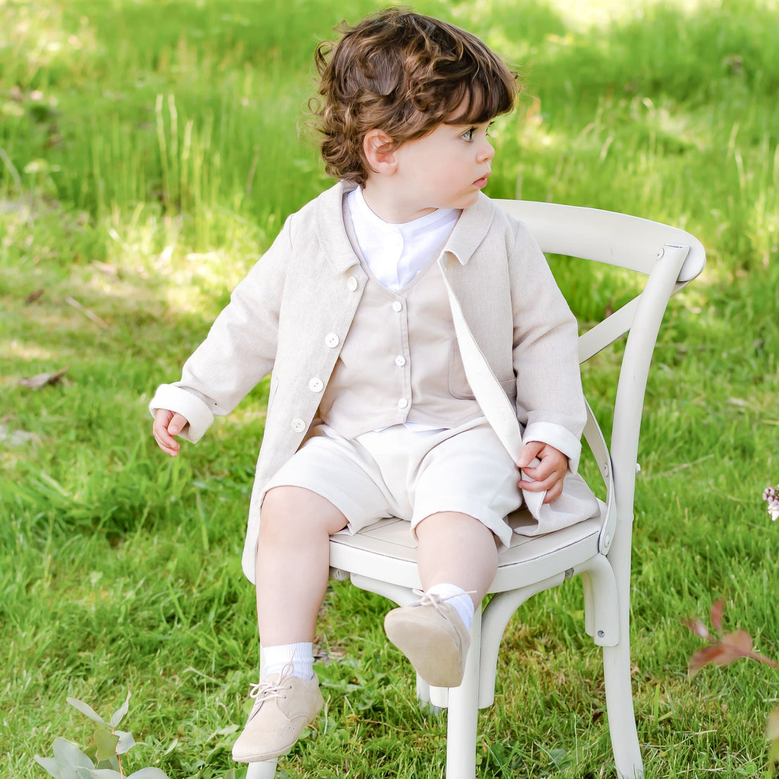 A young child with curly hair sits on a white chair outside, wearing a red linen trench coat over matching shorts and cream-colored shoes. The child looks to the side, surrounded by green grass and a few wildflowers.