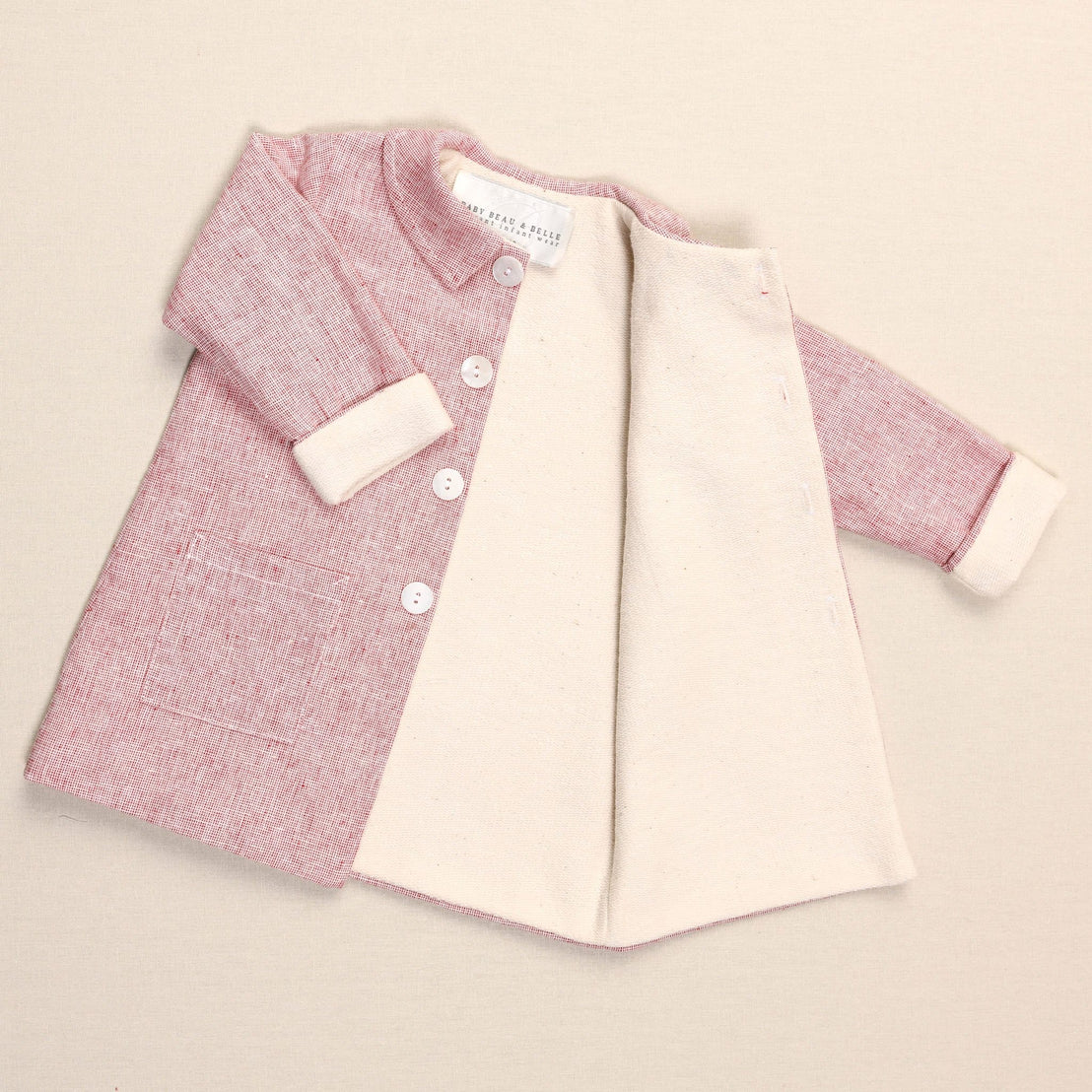 A baby pink and cream handmade coat with buttons and long sleeves is displayed on a beige surface. The coat is open, revealing its cream lining made of soft French terry cotton, and has a front pocket on the left side.