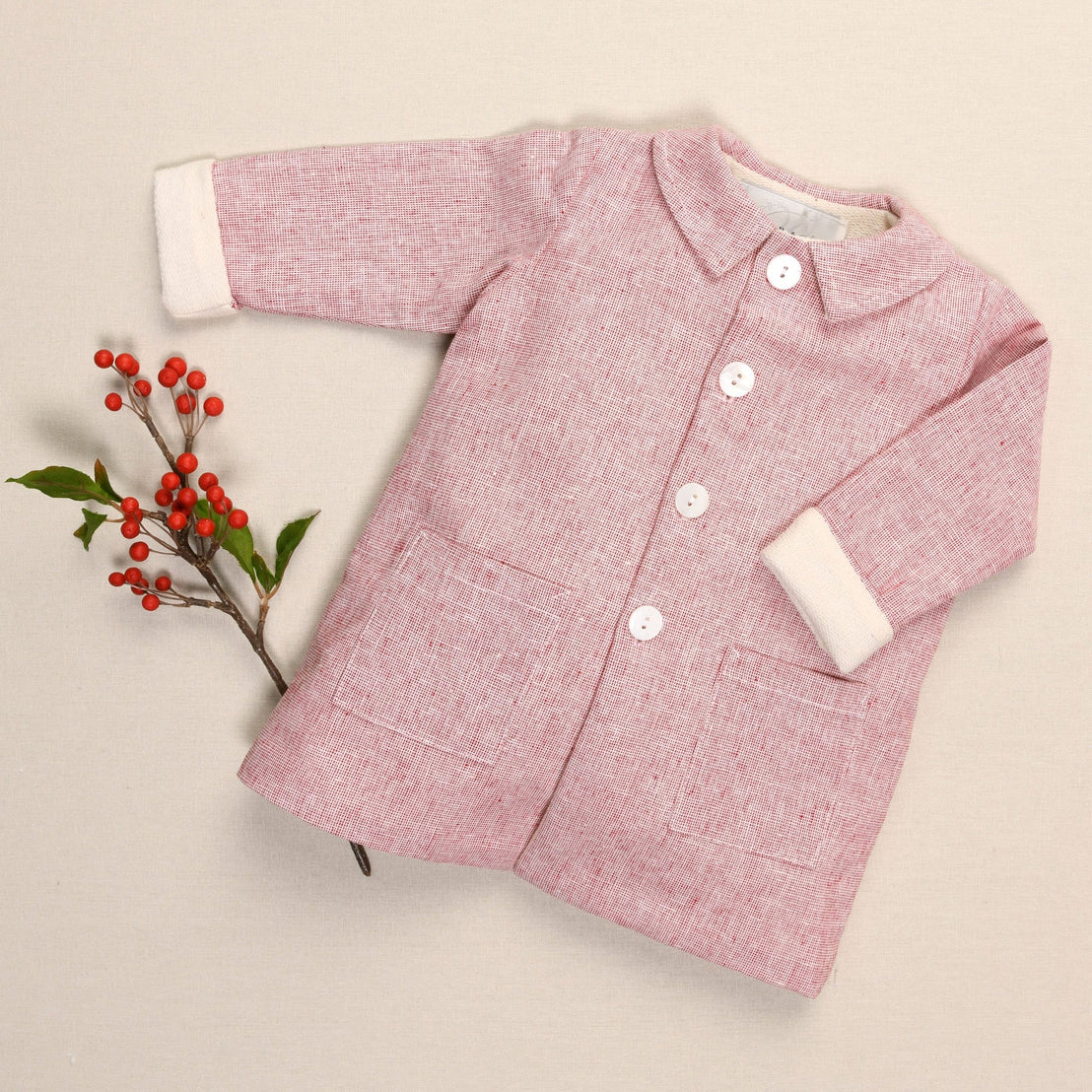 A handmade red linen trench coat for children, adorned with white buttons and two pockets, rests gently against a beige background, accented by a sprig of red berries and green leaves to its left.
