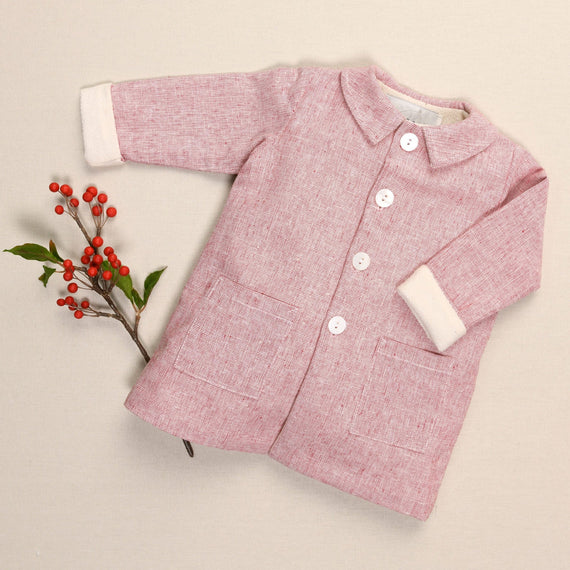 A handmade red linen trench coat for children, adorned with white buttons and two pockets, rests gently against a beige background, accented by a sprig of red berries and green leaves to its left.