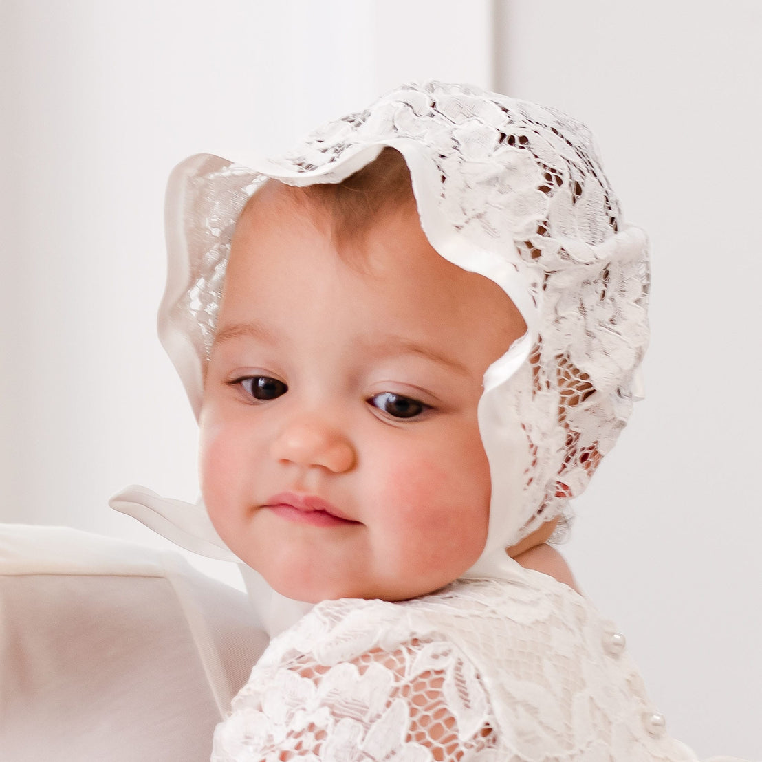 Close-up of a baby girl wearing the Rose Lace Bonnet, made from delicate ivory lace with satin trim. She looks to the side with a soft expression, while the intricate lace details of the bonnet complement her matching gown.
