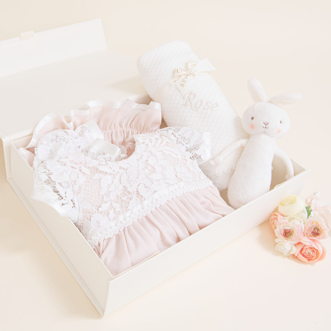 An elegant, traditional Rose Newborn Gift Set containing an heirloom lacy pink dress, a plush white bunny, and a decorative white personalized blanket with "rose" embroidered on it, displayed on a soft peach background