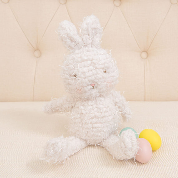 The Shaggy Hoppy Bunny, with its long ears and plush, textured fur that feels just like a dream, sits on a cream-colored couch. Beside the bunny are two small, colorful plastic eggs, one yellow and one pink—ideal for a baby shower gift.