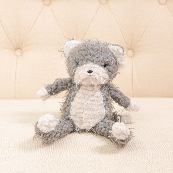 A plush Shaggy Purrty Kitty toy with soft grey and white fur rests on a beige, tufted fabric background. Perfect as a baby shower gift, the cuddly cat has a relaxed pose and a gently worn appearance.