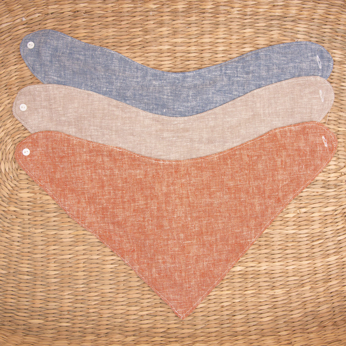 Flat lay photo of all three colors of the Silas Bandana Bib. The bibs are made from linen in three different colors: indigo, sand, and clay.