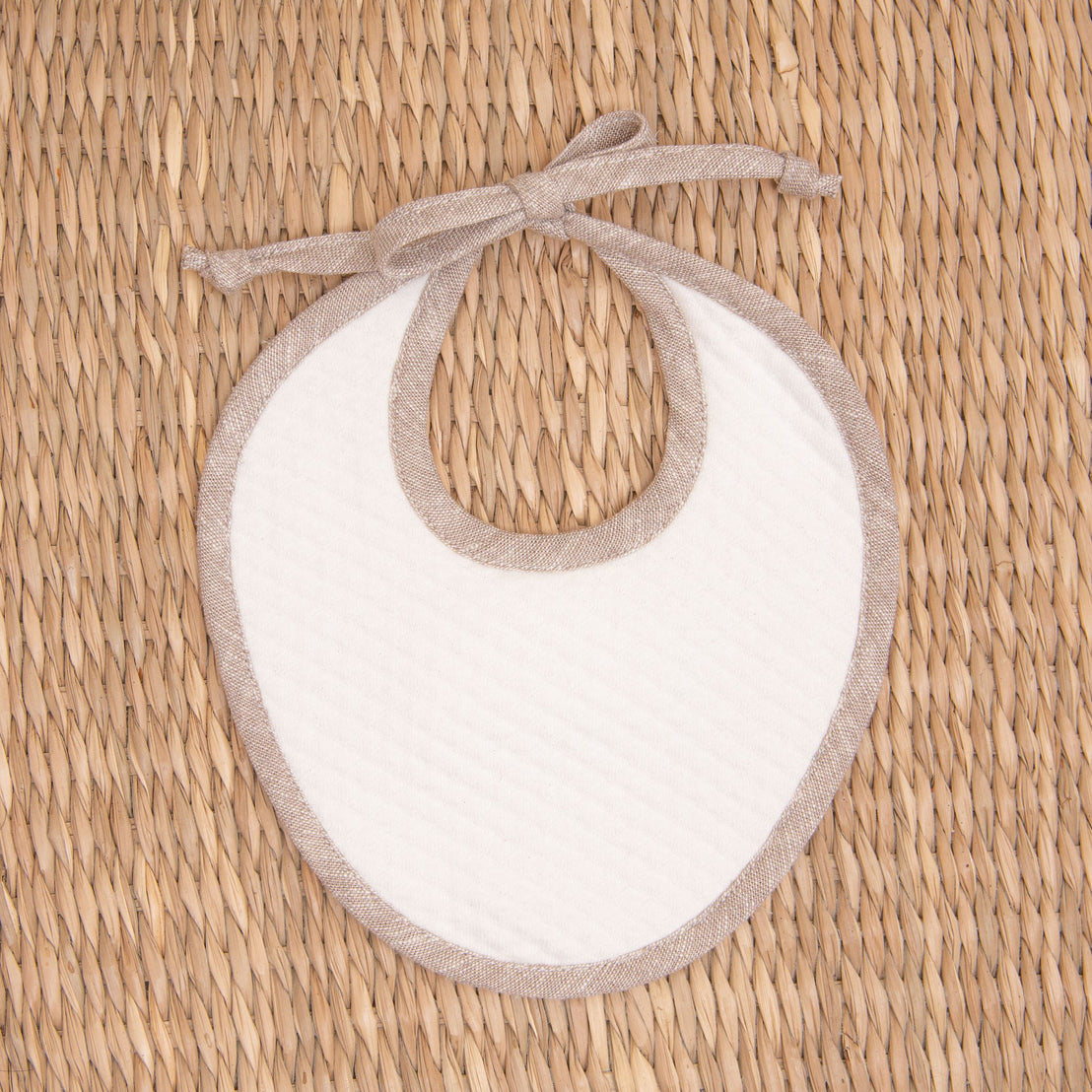 Flat lay photo of the tan Silas Linen Trim Bib. The bib is made from textured cotton in light ivory