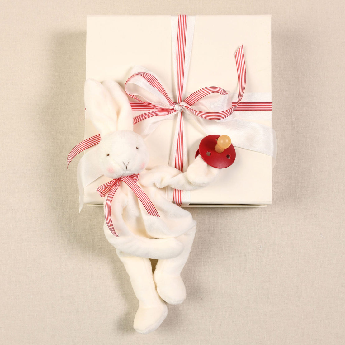 The Silly Bunny Buddy & Pacifier, crafted from soft white velour and decorated with red and white striped ribbons, holds a red wooden rattle. It rests atop a baby gift—a white box tied with a red striped ribbon bow—set against a light beige fabric background.