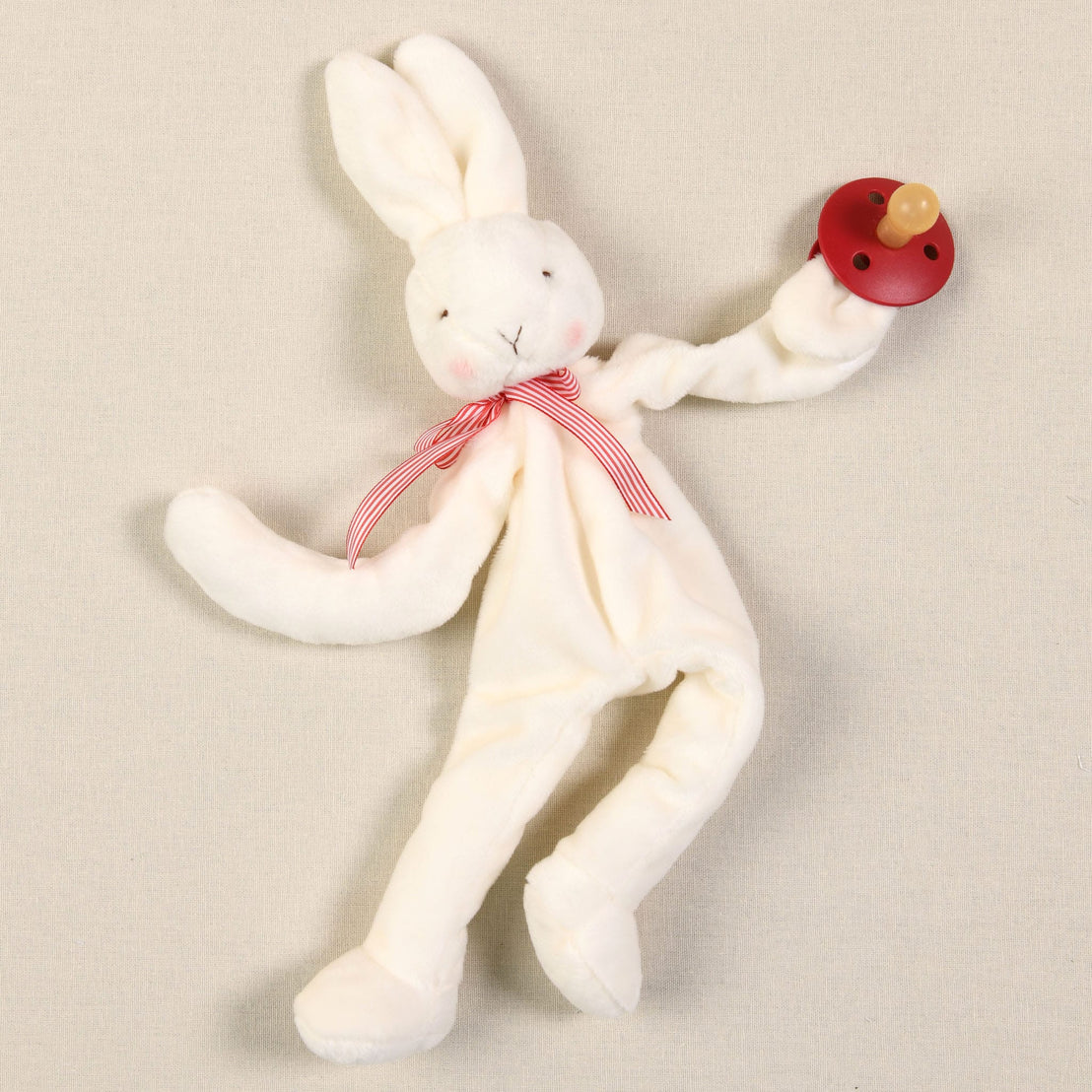 The Silly Bunny Buddy & Pacifier, featuring a plush velour bunny adorned with a red and white striped ribbon around its neck, cradles a wooden toy rattle, making it the perfect baby gift. With long ears and an elegantly embroidered face, this delightful bunny sits on a beige background, ready to become your baby's cherished pacifier holder companion.