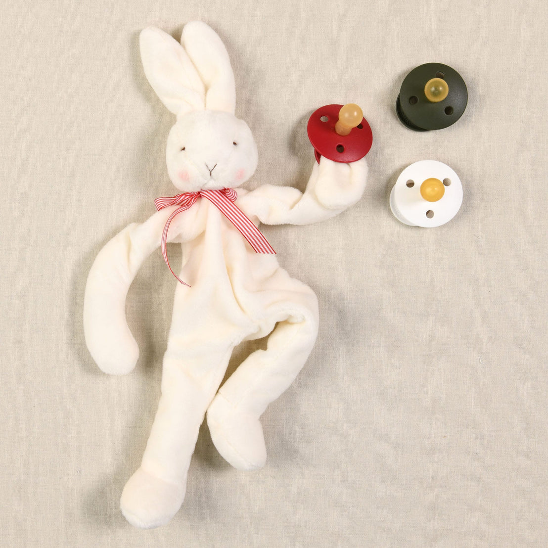 The Silly Bunny Buddy & Pacifier, an ideal gift for babies, rests on a beige surface adorned with a red striped ribbon around its neck. This velour plush rabbit acts as an adorable pacifier holder, clutching a red pacifier while two others—one black and one green—lie nearby.