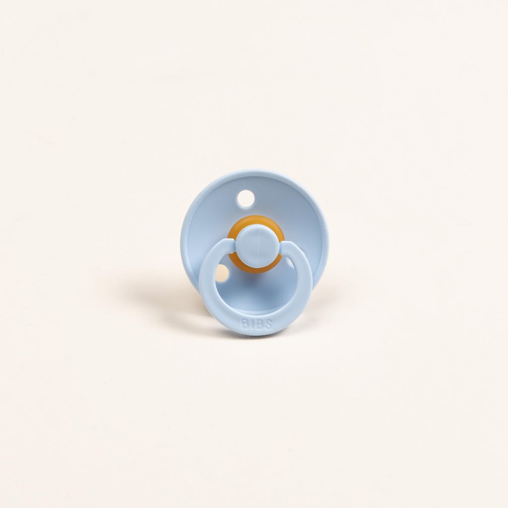 The Silly Bunny Buddy & Pacifier, featuring a light blue color and circular shape with a round, orange rubber nipple, rests against a plain cream-colored background. The base is designed with two small holes and the word "BIBS" engraved on the handle, making it an ideal baby gift when paired with a matching pacifier clip.
