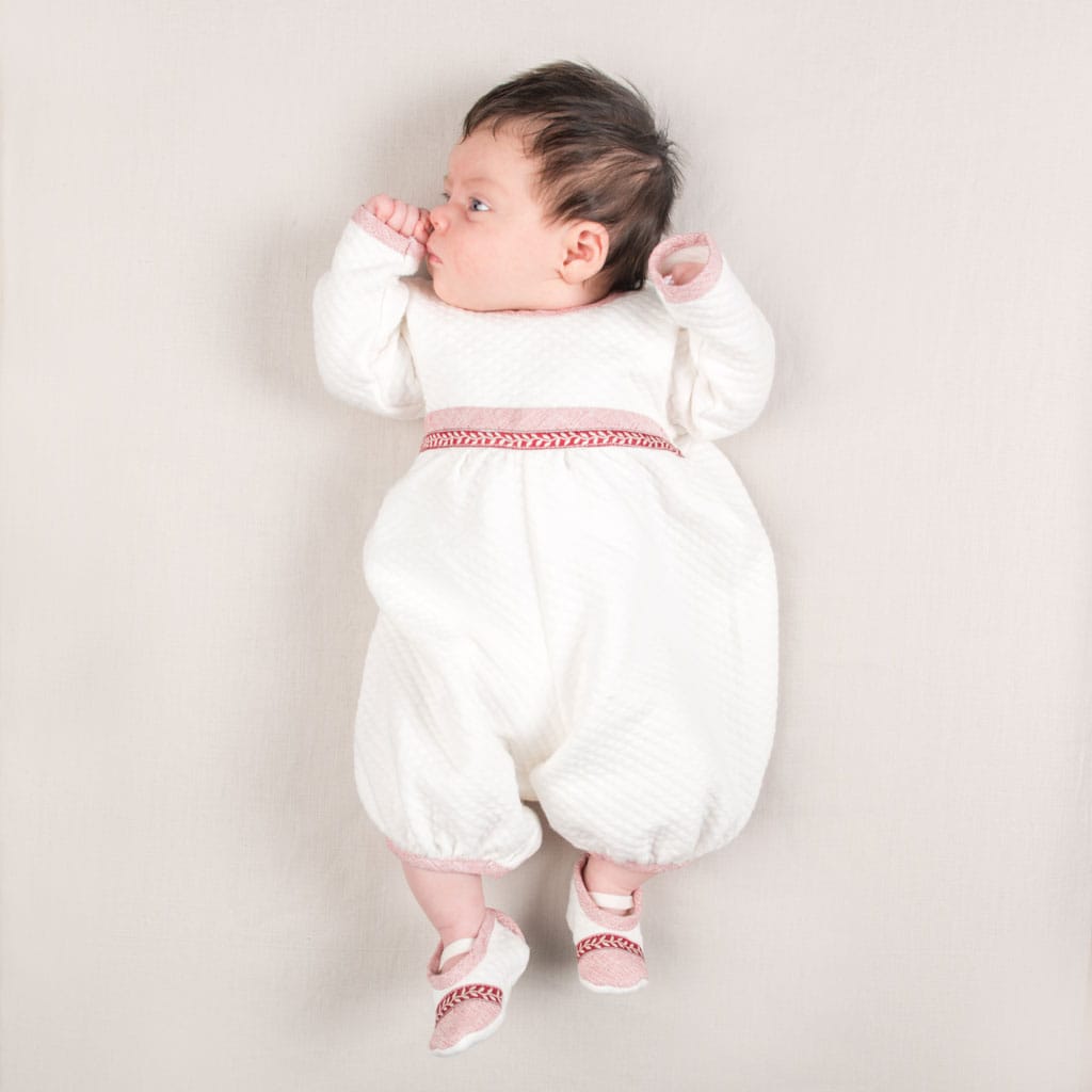 A baby wearing the Sweetheart Newborn Romper, made from 100% quilted cotton with red trim, lies on a light surface. The baby gazes to the side with one hand near their mouth while sporting matching red and white shoes.