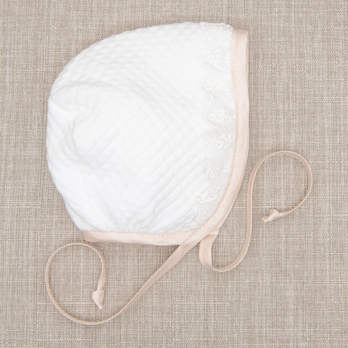 Flat lay of the Tessa Quilt Bonnet laying on a beige fabric surface, highlighting the soft quilted cotton with pink silk trim and delicate ivory floral lace accents.