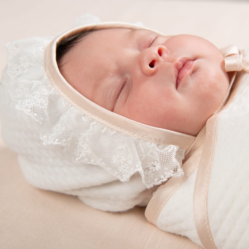 Close-up of a newborn baby peacefully sleeping while wearing the Tessa Quilted Bonnet. The bonnet features a soft ivory quilted fabric with delicate lace trim and satin binding.