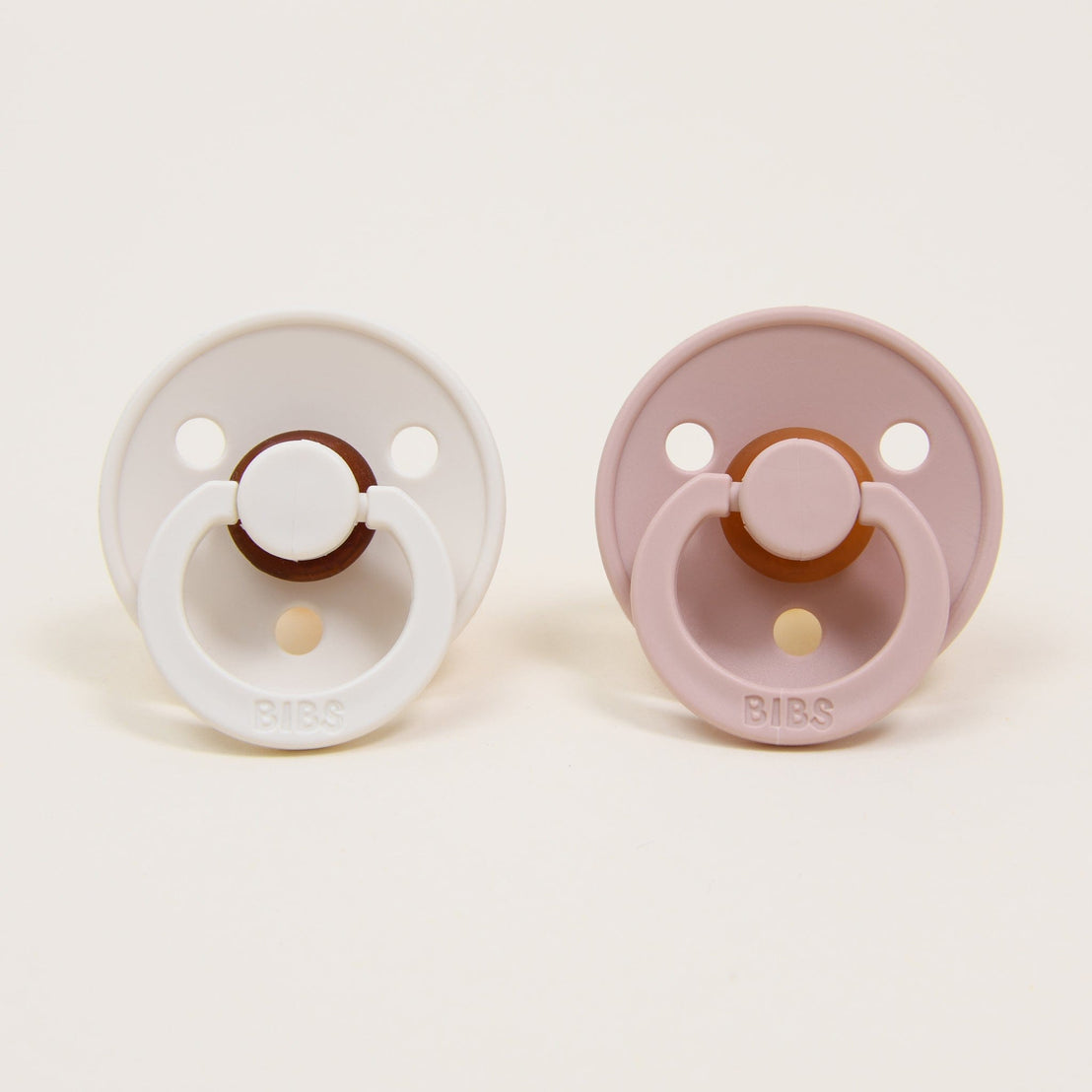 The Tessa Pacifier Set, featuring two baby pacifiers crafted with natural rubber, is displayed side by side on a neutral background. The left one is in ivory and the right in blush, each showcasing a circular base and a round handle embossed with "BIBS," a trusted Danish brand known for helping babies self-soothe.