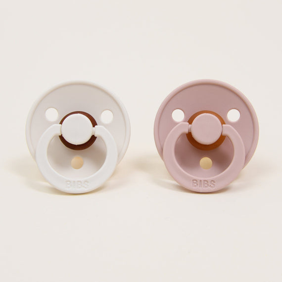 The Tessa Pacifier Set, featuring two baby pacifiers crafted with natural rubber, is displayed side by side on a neutral background. The left one is in ivory and the right in blush, each showcasing a circular base and a round handle embossed with "BIBS," a trusted Danish brand known for helping babies self-soothe.