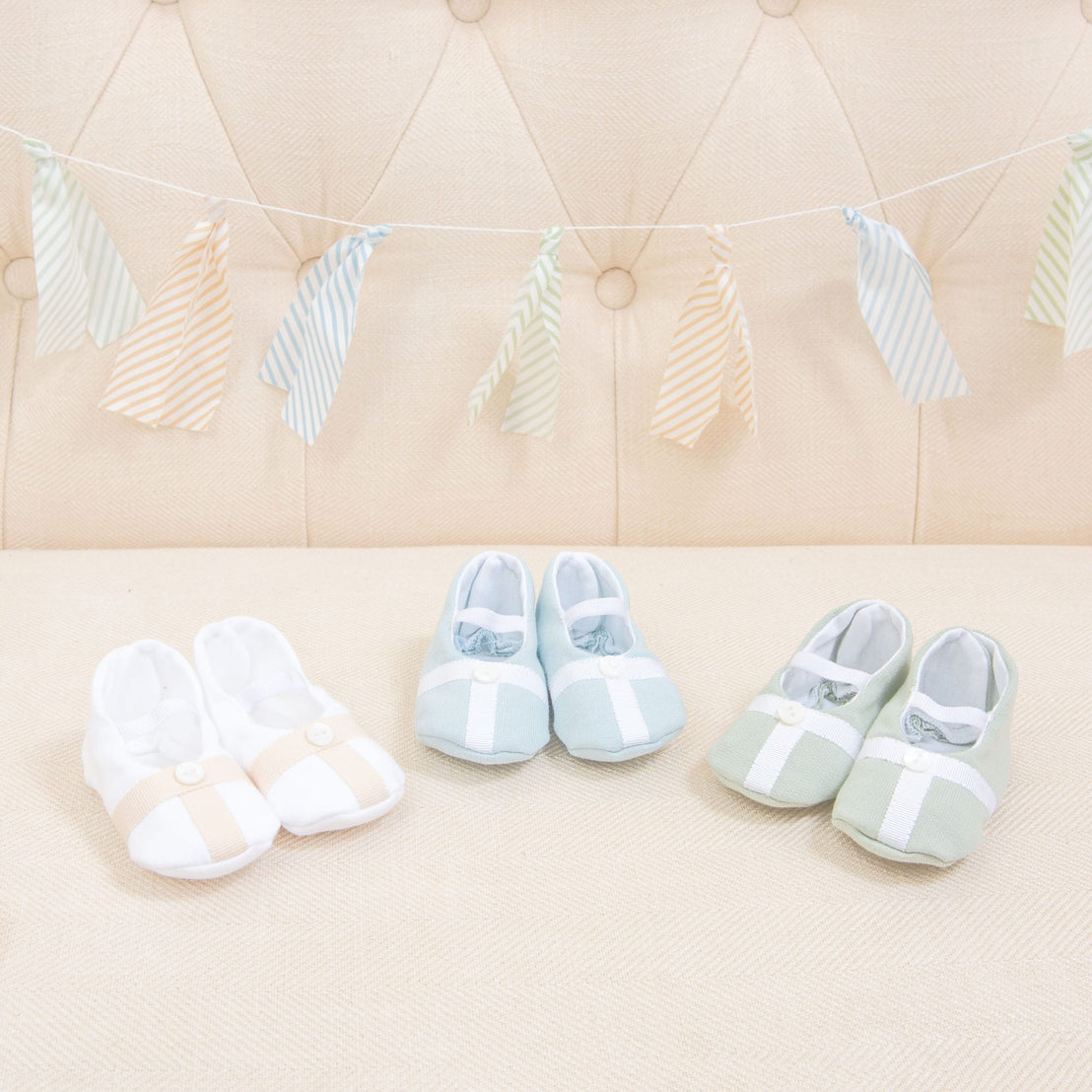 Flat lay photo showing the top detail of three pairs of Theodore Booties, including blue, green, and a white/tan color. The booties are made from a 100% French Terry Cotton with a Grosgrain Trim (with button detail) and soft elastic strap.
