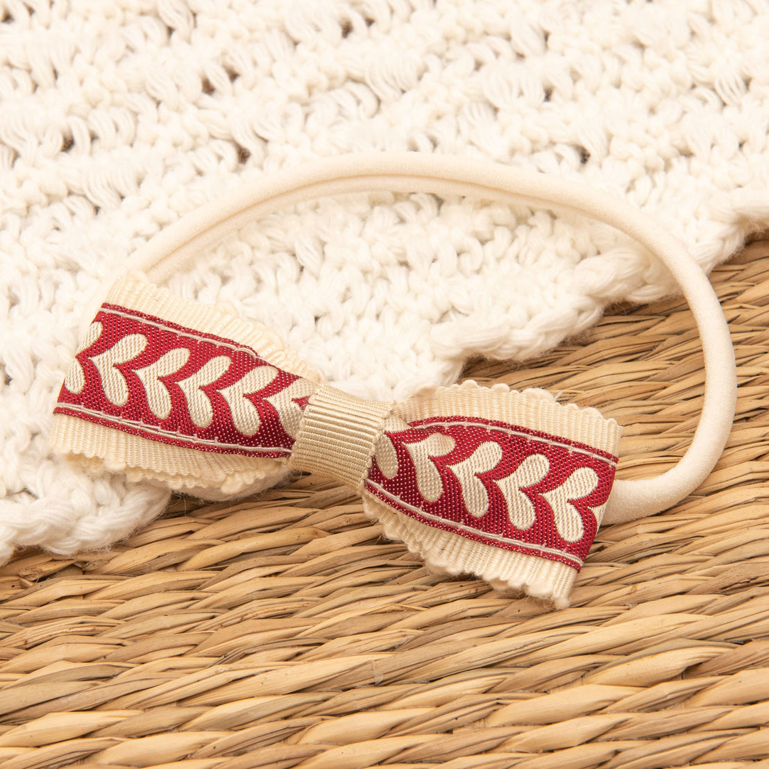 A beige headband adorned with a red and beige leaf-patterned bow rests on a woven straw surface, complemented by textured white quilted cotton fabric—an elegant component of the Valentina Gift Set, now available at a 10% discount.