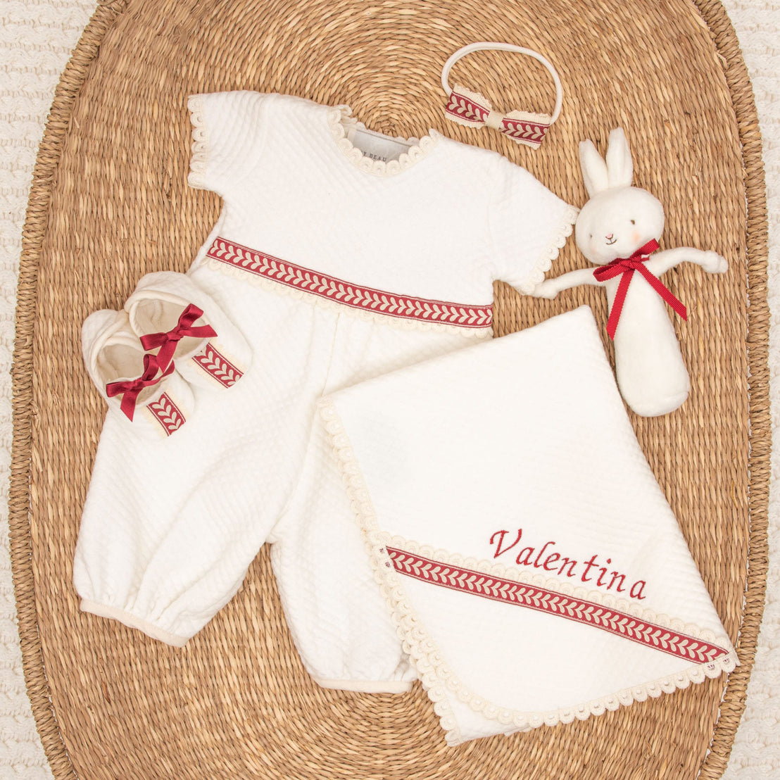 The Valentina Gift Set - Save 10% features a beautifully arranged baby gift on a woven mat, including the delightful Valentina romper with red accents, a coordinating headband, matching shoes, an adorable plush bunny toy, and a personalized quilted cotton blanket embroidered with the name "Valentina" in red.