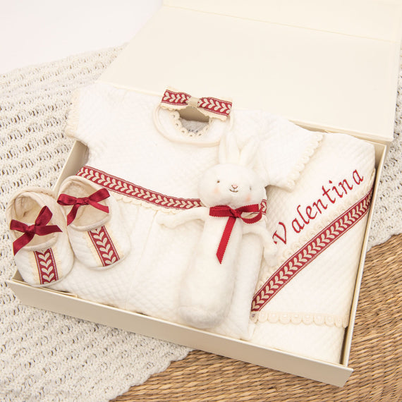 The Valentina Gift Set, offering a 10% savings, features a beautifully organized selection of baby gifts, including a white dress, plush bunny, matching booties, and a personalized towel embroidered with "Valentina." In addition to these items is a cozy romper adorned with red ribbon accents. Everything is elegantly displayed in an open box on a knitted and woven surface.