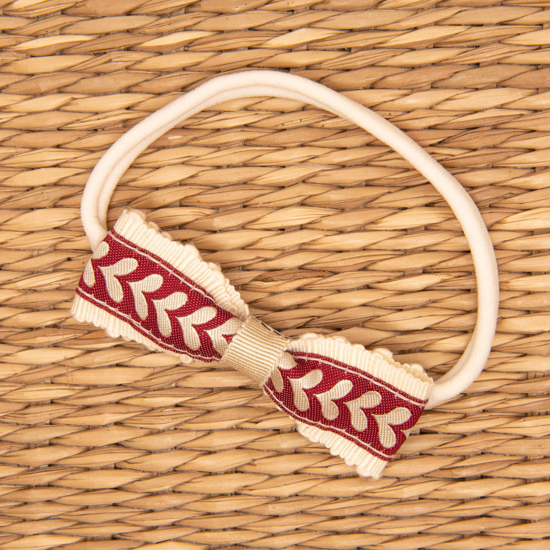 The Valentina Headband, featuring a red and beige patterned bow with a symmetrical leaf design, rests on a woven straw surface. Its handmade circular cream-colored elastic band is perfect for complementing any holiday outfit.
