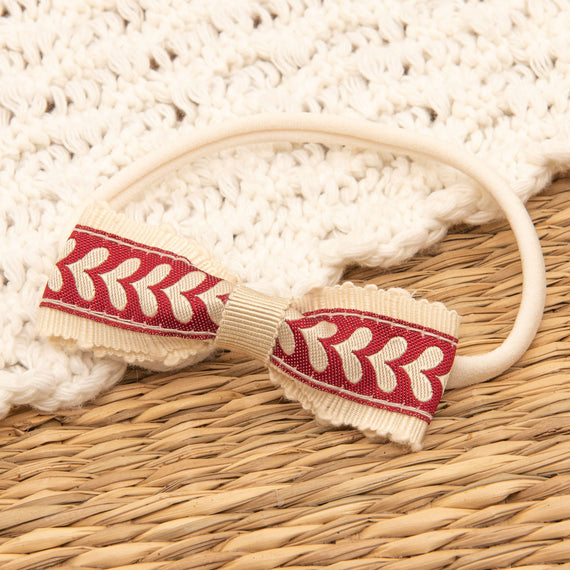 The Valentina Headband, featuring a handmade beige band with a red and white patterned bow, rests on a woven surface. It's an ideal accessory for adding charm to any holiday outfit and comes with textured white fabric.