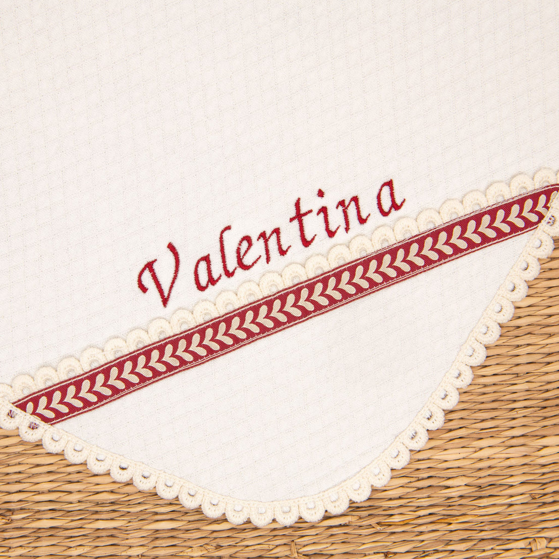 The "Valentina Holiday Personalized Blanket" features an ivory quilted cotton fabric with scalloped edges, beautifully embroidered with the name "Valentina" in red script. A decorative red and white woven border, reminiscent of a holiday ribbon, accentuates the lower edge, enhancing its festive charm. This handmade receiving blanket is elegantly displayed on a woven surface.