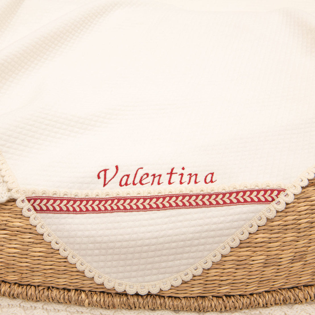 The Valentina Holiday Personalized Blanket, a creamy white handmade creation crafted from ivory quilted cotton, is elegantly draped over a woven basket. It features the embroidered name "Valentina" in red and is adorned with a decorative red and white trim along the edge.