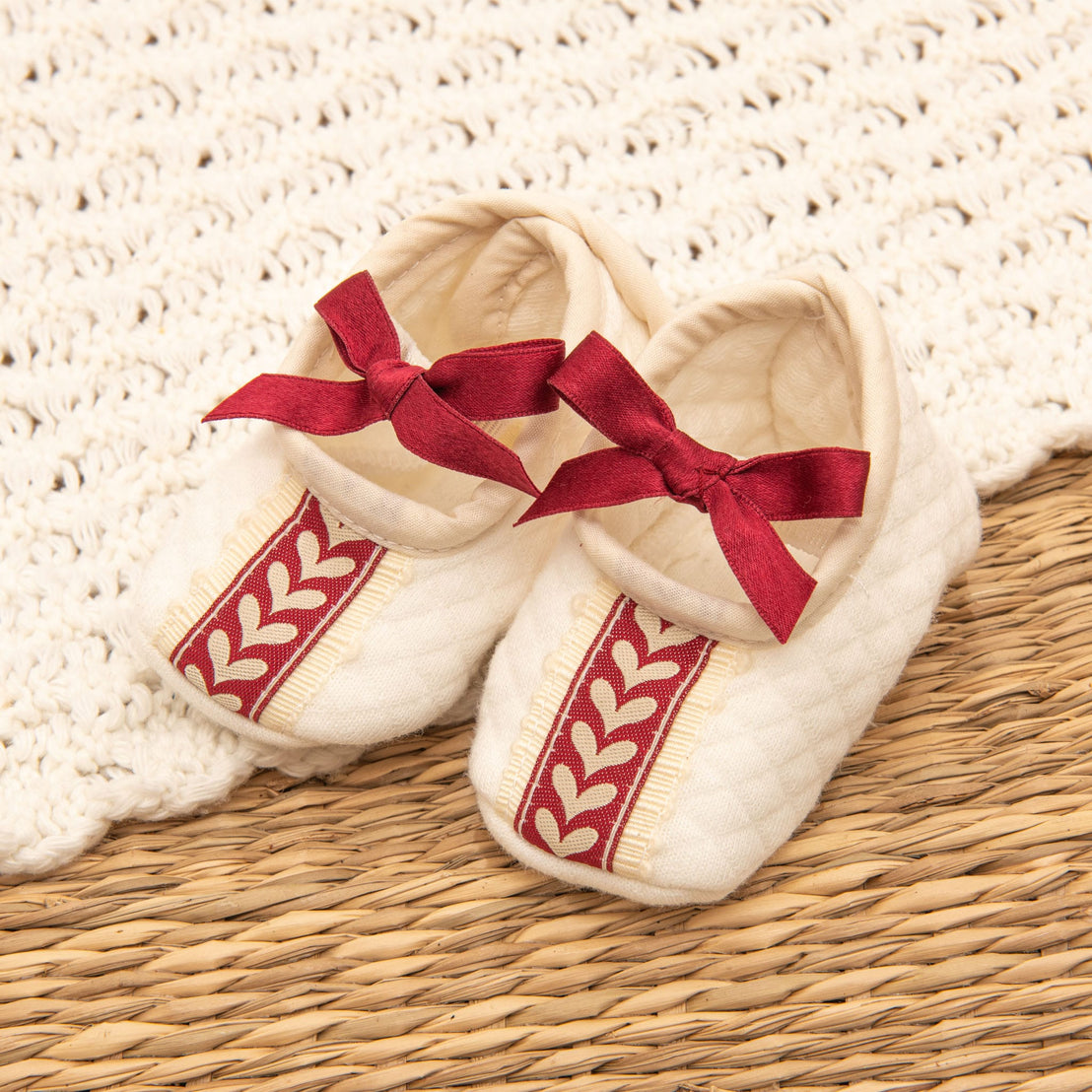 The Valentina Quilted Booties are cream-colored baby booties adorned with red ribbons and decorative red and cream leaf-patterned stripes. Ideal for a newborn photo shoot, they rest on a tan woven surface with a cream crochet backdrop, making them perfect for your baby's first Christmas outfit.