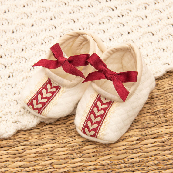 The Valentina Quilted Booties are cream-colored baby booties adorned with red ribbons and decorative red and cream leaf-patterned stripes. Ideal for a newborn photo shoot, they rest on a tan woven surface with a cream crochet backdrop, making them perfect for your baby's first Christmas outfit.
