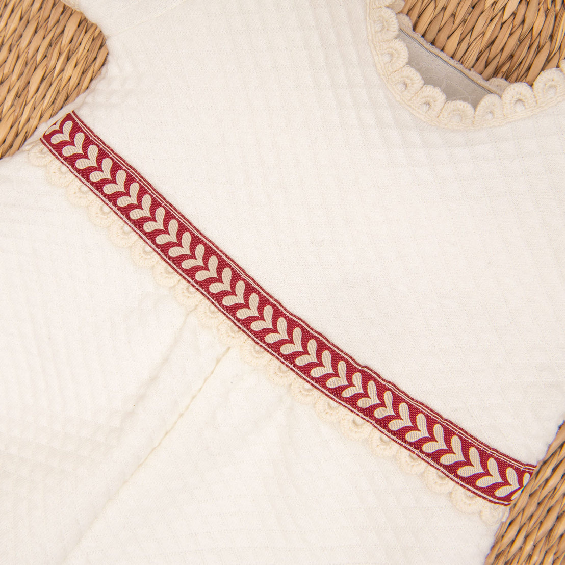 Close-up of the Valentina Romper, showcasing its cream-colored textured fabric, adorned with a decorative red and beige trim. The intricate leaf pattern with scalloped edges adds charm, making it perfect for a newborn Christmas outfit as it rests on a woven surface.