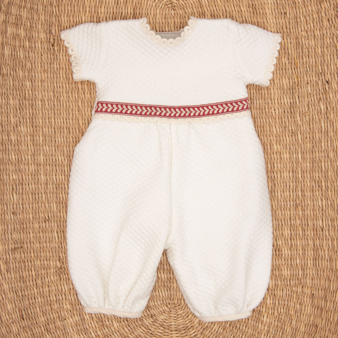The Valentina Romper is a white infant outfit with short sleeves and textured fabric, featuring delicate lace edging. Ideal for a newborn Christmas ensemble, it has a red and white herringbone patterned band at the waist. The romper is displayed flat against a woven straw mat background.
