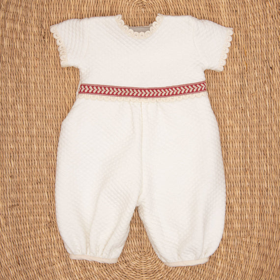 The Valentina Romper is a white infant outfit with short sleeves and textured fabric, featuring delicate lace edging. Ideal for a newborn Christmas ensemble, it has a red and white herringbone patterned band at the waist. The romper is displayed flat against a woven straw mat background.