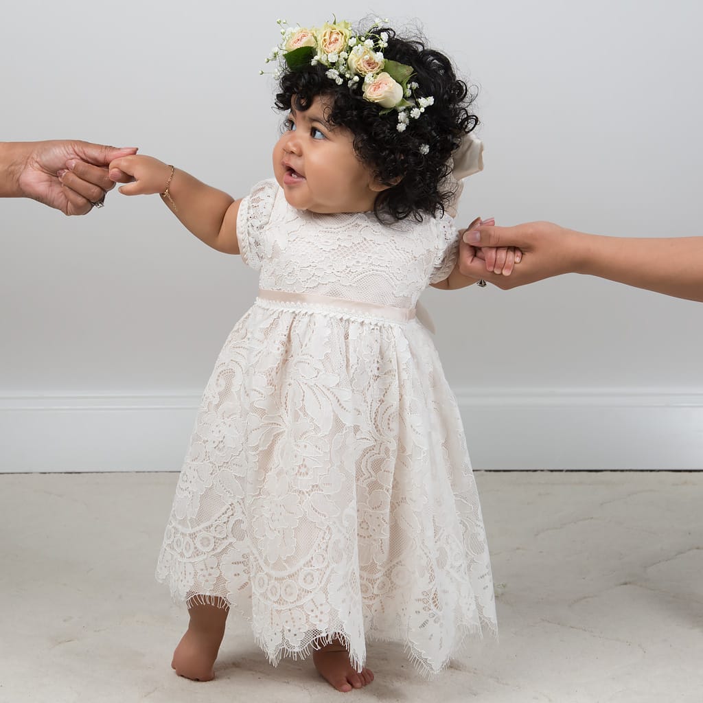A baby in a Victoria Puff Sleeve Dress and floral headband stands barefoot, holding hands with two adults on either side against a plain background. She has curly hair and appears to be smiling slightly.