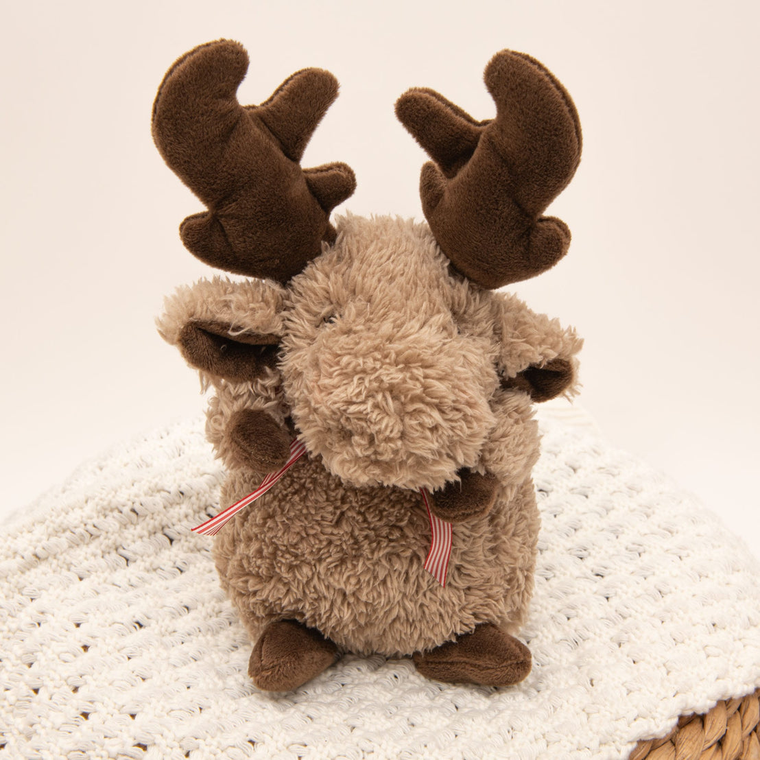 A Wee Reindeer with a light brown body, soft fur, and dark brown antlers sits on a white textured blanket. It features a red and white striped ribbon around its neck, making it perfect for snuggles.