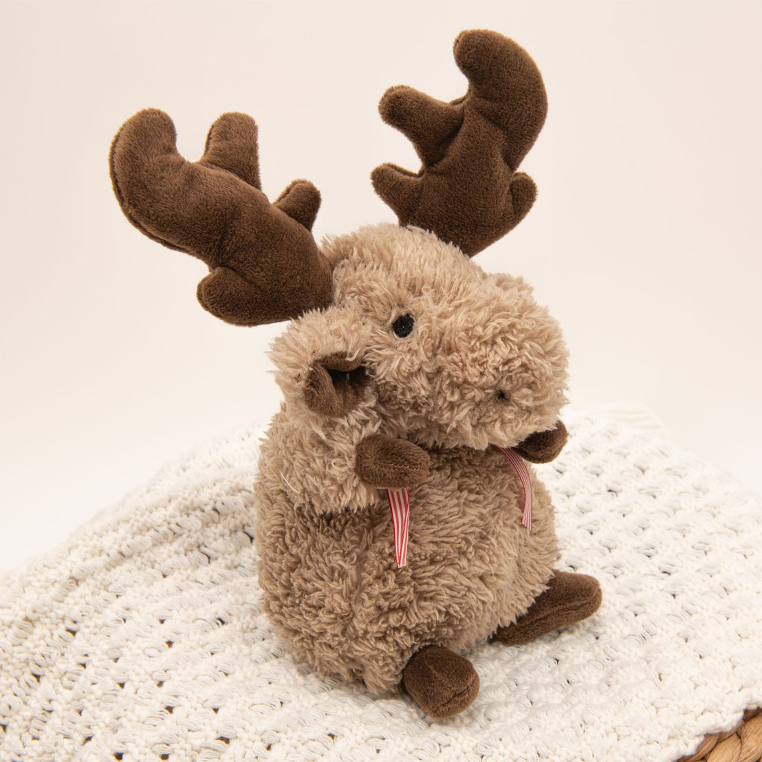 A plush Wee Reindeer toy with soft fur and dark brown antlers rests on a white crocheted blanket. Its light brown body is adorned with small, round ears and a tiny pink ribbon around the neck. Safe for babies and incredibly cute, it stands out against the simple, light-colored background.