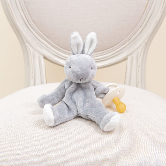 A Wee Silly Bunny Pacifier Holder, plush and gray, sits on a cream-colored upholstered chair. Designed to be safe for babies, this adorable rabbit securely holds a white and yellow pacifier.