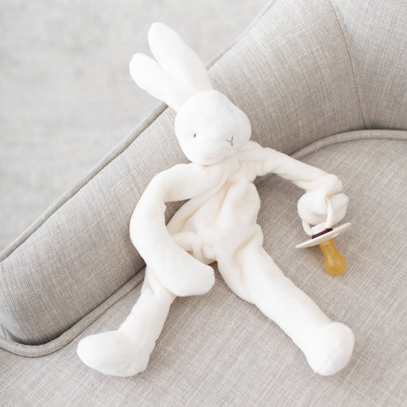 A plush toy named the "White Silly Bunny Buddy | Pacifier Holder" is resting on a light gray couch. The bunny, with its floppy ears and simple facial features, holds a pacifier attached to its clip, creating a cozy and gentle atmosphere—an ideal gift for a baby.