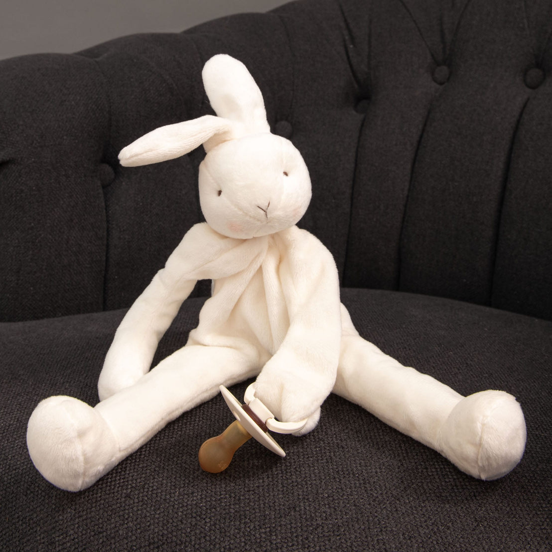 The White Silly Bunny Buddy | Pacifier Holder, ideal as a christening gift, rests on a dark gray couch, holding a baby's pacifier in its paw. Its soft white fur gently contrasts with the background while ensuring it's completely safe for babies.