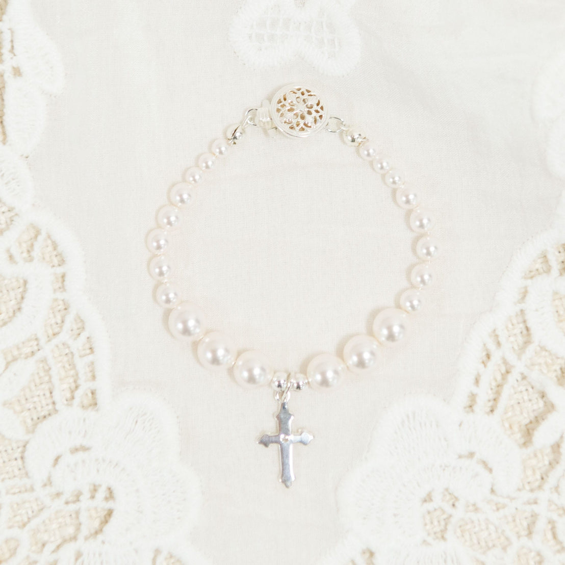 A flat lay photo baby jewelry: the white luster pearl bracelet with a silver cross in the middle. Photographed on top of the Adeline Christening Gown.