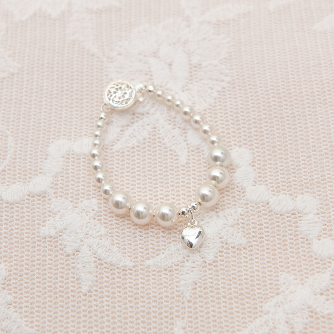 A White Luster Pearl bracelet with an upscale Silver Heart charm, displayed on a delicate pink lace background.