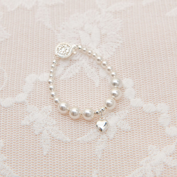 A White Luster Pearl bracelet with an upscale Silver Heart charm, displayed on a delicate pink lace background.