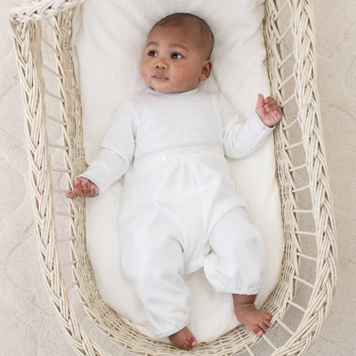 Baby wearing the Harrison Long Sleeve Romper in a natural basket, showcasing its elegant white quilted fabric and delicate lace details.
